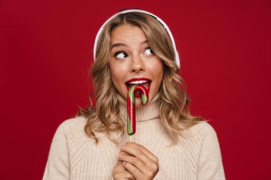 Kırmızı arka planda izole edilmiş şeker kamışlı Noel Baba şapkası takan sevimli, neşeli genç bir kadın.