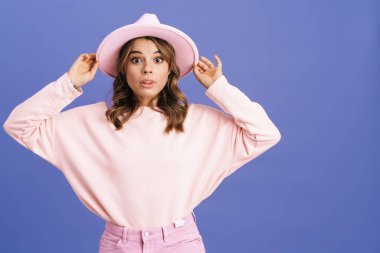 Şok olmuş şapkalı çekici kız mor arka planda izole edilmiş kameraya şaşırdığını ifade ediyor.