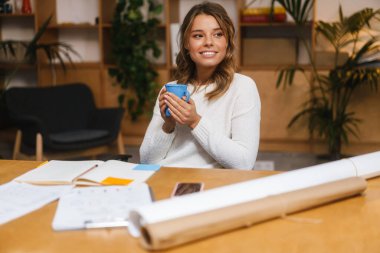 Ofis masasında oturup kahve içerken belgelerle çalışan güzel, gülümseyen genç bir girişimci.