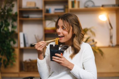 Gülümseyen genç girişimci kadın ofis masasında oturmuş erişte yerken öğle yemeği yiyor.