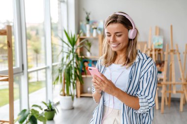Güzel, mutlu, genç bir tasarımcının portresi. Yaratıcı ofiste kulaklık takıyor, cep telefonu tutuyor.