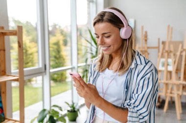 Güzel, mutlu, genç bir tasarımcının portresi. Yaratıcı ofiste kulaklık takıyor, cep telefonu tutuyor.