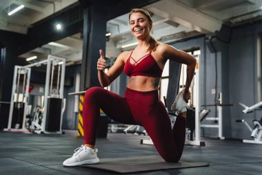 Gülümseyen sporcu kadın spor salonunda paspas üzerinde çalışırken başparmağını gösteriyor.