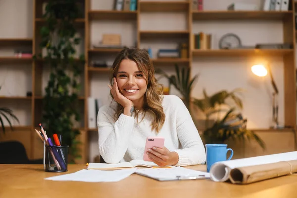 Smiling attractive young woman entrepreneur using mobile phone while working with documents at the office desk