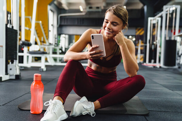Счастливая спортсменка с мобильным телефоном и улыбкой сидя на коврике в тренажерном зале