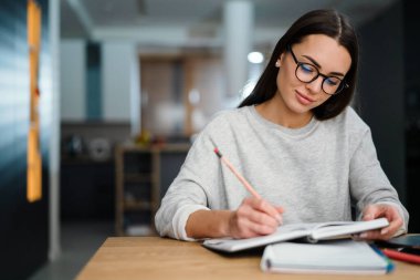 Gözlüklü genç bir kadın evde çalışırken notlar yazıyor.