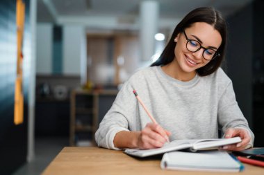 Gözlüklü mutlu genç kadın evde çalışırken notlar yazıyor.