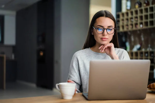 眼鏡をかけながら自宅でノートパソコンと仕事をしながらコーヒーを飲む若い女性 — ストック写真