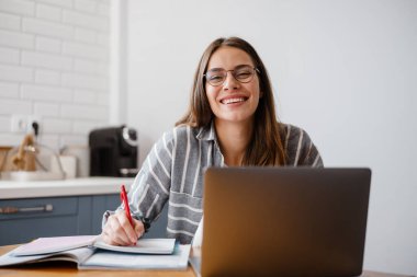 Evdeki masada oturmuş laptopla ödev yapan neşeli güzel bir kadın.