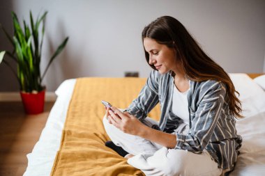 Evdeki yatakta otururken cep telefonu kullanan mutlu esmer kadın.