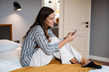 Evdeki yatakta otururken cep telefonu kullanan mutlu esmer kadın.