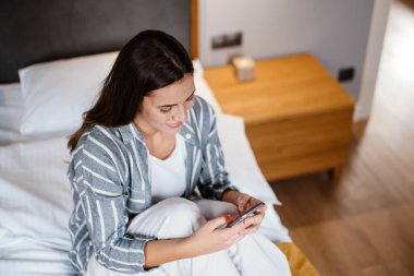 Evdeki yatakta otururken cep telefonu kullanan mutlu esmer kadın.