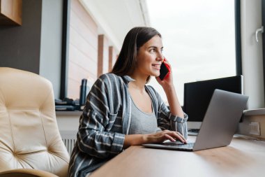 Evde dizüstü bilgisayarla çalışırken cep telefonuyla konuşan hoş, mutlu bir kadın.