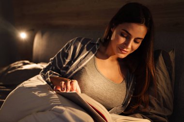 Evde yatakta uzanmış kitap okurken gülümseyen ve mutlu bir kadın.