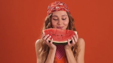 Kafasında bandana olan, bir dilim karpuz gösteren ve turuncu stüdyoda ısıran kıvırcık saçlı bir kadın.