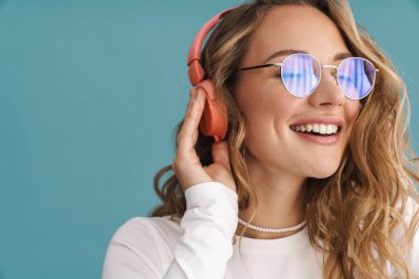 Mavi duvarda kablosuz kulaklıkla müzik dinleyen, gözlüklü, gülümseyen sarışın bir kadın.