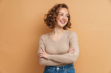 Kızıl saçlı genç kadın kollarını kavuşturup gülümsüyor. Bej arka planda izole bir şekilde duruyor.