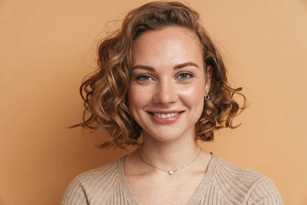 Young ginger woman with wavy hair smiling and looking at camera isolated over beige background