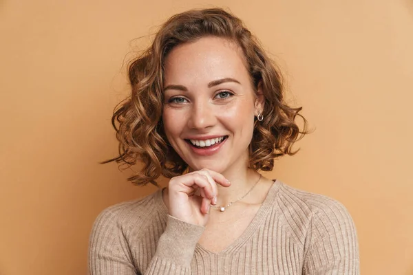 Young ginger woman with wavy hair smiling and looking at camera isolated over beige background