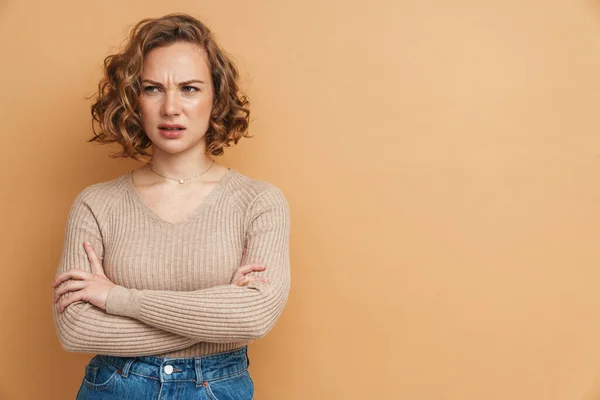 Displeased ginger woman frowning while posing with arms crossed isolated over beige background