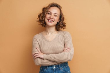 Kızıl saçlı genç kadın kollarını kavuşturup gülümsüyor. Bej arka planda izole bir şekilde duruyor.