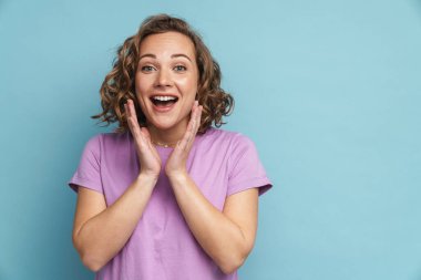 Dalgalı saçlı ve gülümseyen kızıl saçlı genç kadın mavi arka planda izole edilmiş kameraya bakıyor.