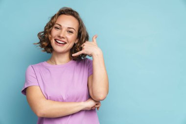 Dalgalı saçlı, gülümseyen ve el hareketi yapan genç kızıl kadın mavi arka planda izole edilmiş.