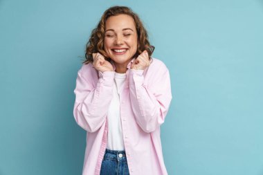 Mavi arka planda izole edilmiş yumruklarını sıkarken dalgalı saçlı genç kızıl kadın gülümsüyor.