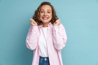 Mavi arka planda izole edilmiş yumruklarını sıkarken dalgalı saçlı genç kızıl kadın gülümsüyor.