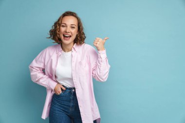 Dalgalı saçlı, gülümseyen ve işaret eden genç kızıl kadın mavi arka planda soyutlanmış.