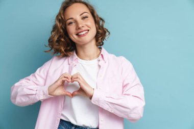 Dalgalı saçlı, gülümseyen ve kalp hareketi gösteren genç kızıl kadın mavi arka planda izole edilmiş.