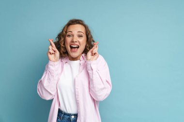 Kızıl saçlı genç bir kadın, mavi arka planda şans dilemek için parmaklarını çapraz tutarak gülüyor.