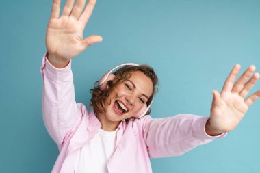 Genç kızıl kadın mavi arka planda izole edilmiş kulaklıklarla müzik dinlerken dans ediyor.