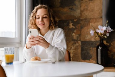 Beyaz sarışın kadın cep telefonu kullanıyor ve kahvaltısını evde yapıyor.