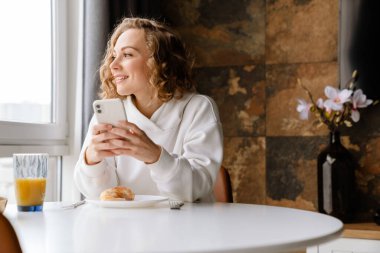 Beyaz sarışın kadın cep telefonu kullanıyor ve kahvaltısını evde yapıyor.