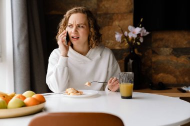 Evde kahvaltı ederken cep telefonuyla konuşan genç beyaz bir kadın.