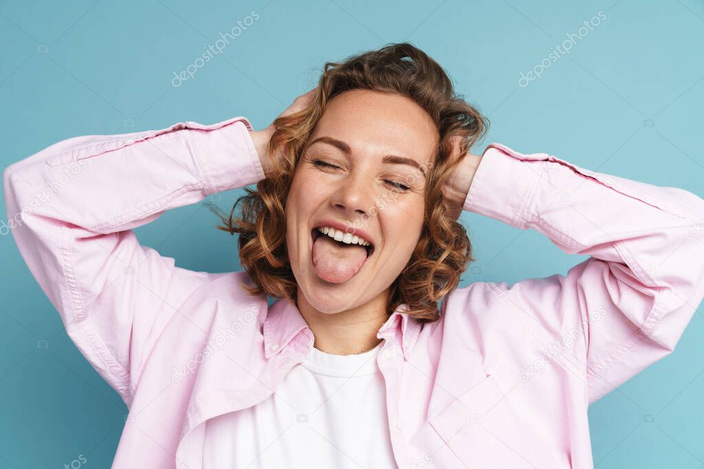 Young ginger woman showing her tongue while holding her head isolated over blue background