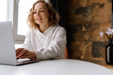 Beyaz sarışın kadın evde otururken dizüstü bilgisayarında gülümsüyor ve daktilo yazıyor.