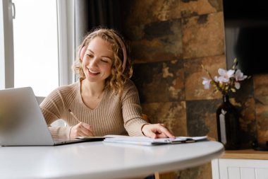 Kulaklıklı genç bir kadın evde dizüstü bilgisayarla çalışırken notlar yazıyor.