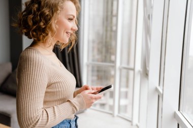 Evde cep telefonu kullanırken gülümseyen genç sarışın kadın.