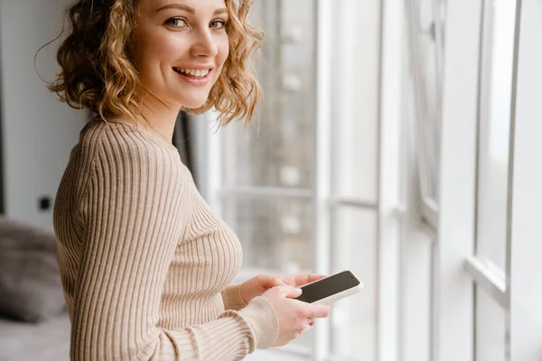 Junge Blonde Frau Lächelt Während Sie Hause Ihr Handy Benutzt — Stockfoto