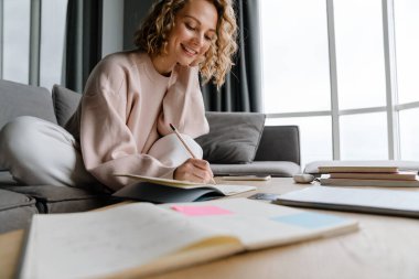 Genç beyaz kadın gülümsüyor ve evdeki kanepede otururken notlar yazıyor.