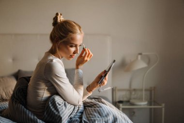 Beyaz sarışın kadın evde yatağında otururken kaşlarını yoluyor.