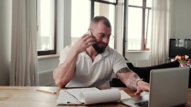 A pleased bearded man wearing eyeglasses is using his laptop while talking on the smartphone sitting at home