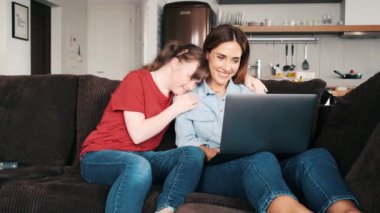 A beautiful woman is looking to her laptop while her daughter with down syndrome is hugging her sitting on the couch at home