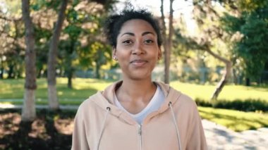 Gülümseyen Afrikalı Amerikalı bir kadın parkta dikilirken kameraya bakıyor.