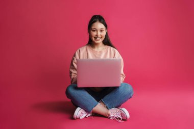 Asyalı gülümseyen kadın mor duvarın üzerinde tek başına otururken dizüstü bilgisayar kullanıyor.