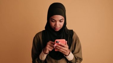 Gülümseyen bir Arap kadın, bej arka planda tek başına duran akıllı telefonunu kullanıyor.