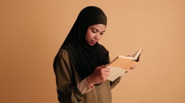 Memnun bir Arap kadın elinde bir kitap tutuyor ve gülümsüyor. Bej arka planda tek başına duruyor.