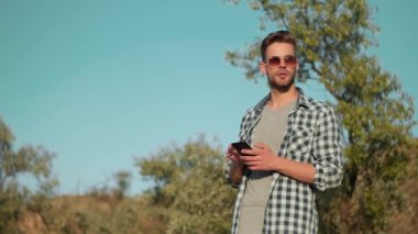 Güneş gözlüğü takan çekici bir adam yazın dışarıda yürürken akıllı telefonunu kullanıyor.